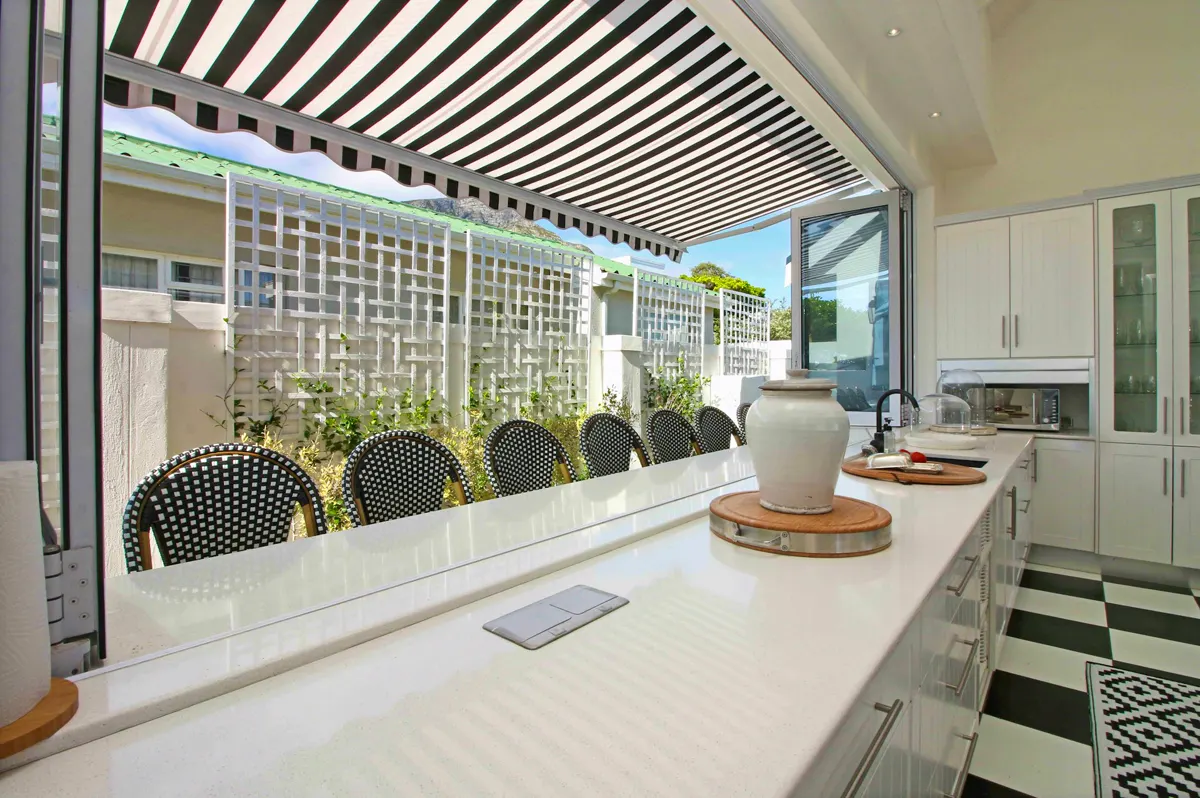 The kitchen at Voelklip Villa opening through a wide serving hatch onto a shaded outdoor dining terrace with seating for a large group